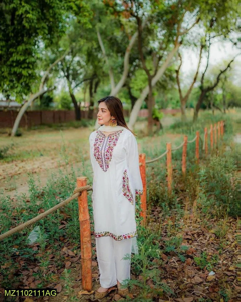 Woman in a white embroidered outfit standing in a natural setting with trees and a wooden fence.