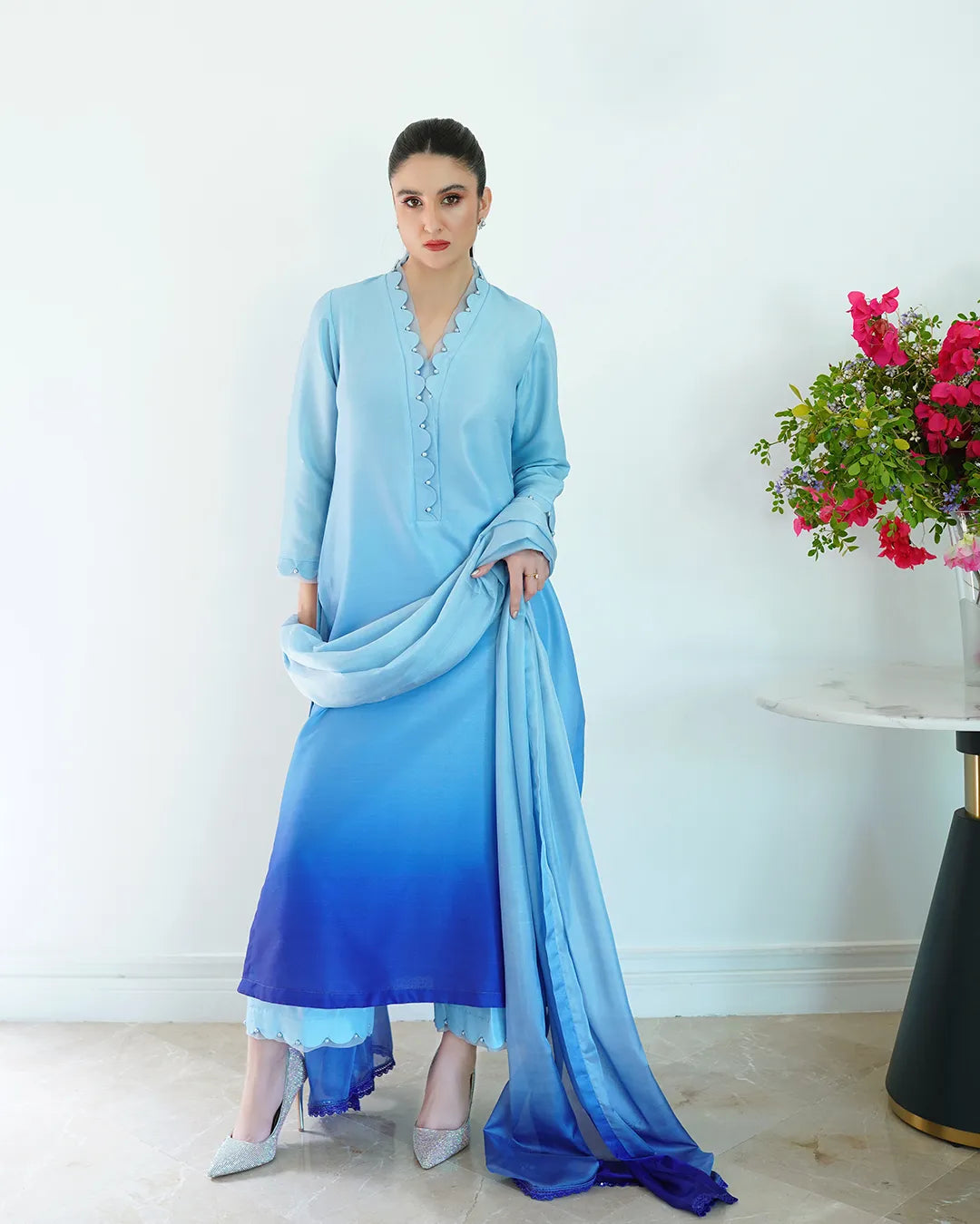 Woman in a light blue outfit with a matching scarf, standing next to a table with flowers.