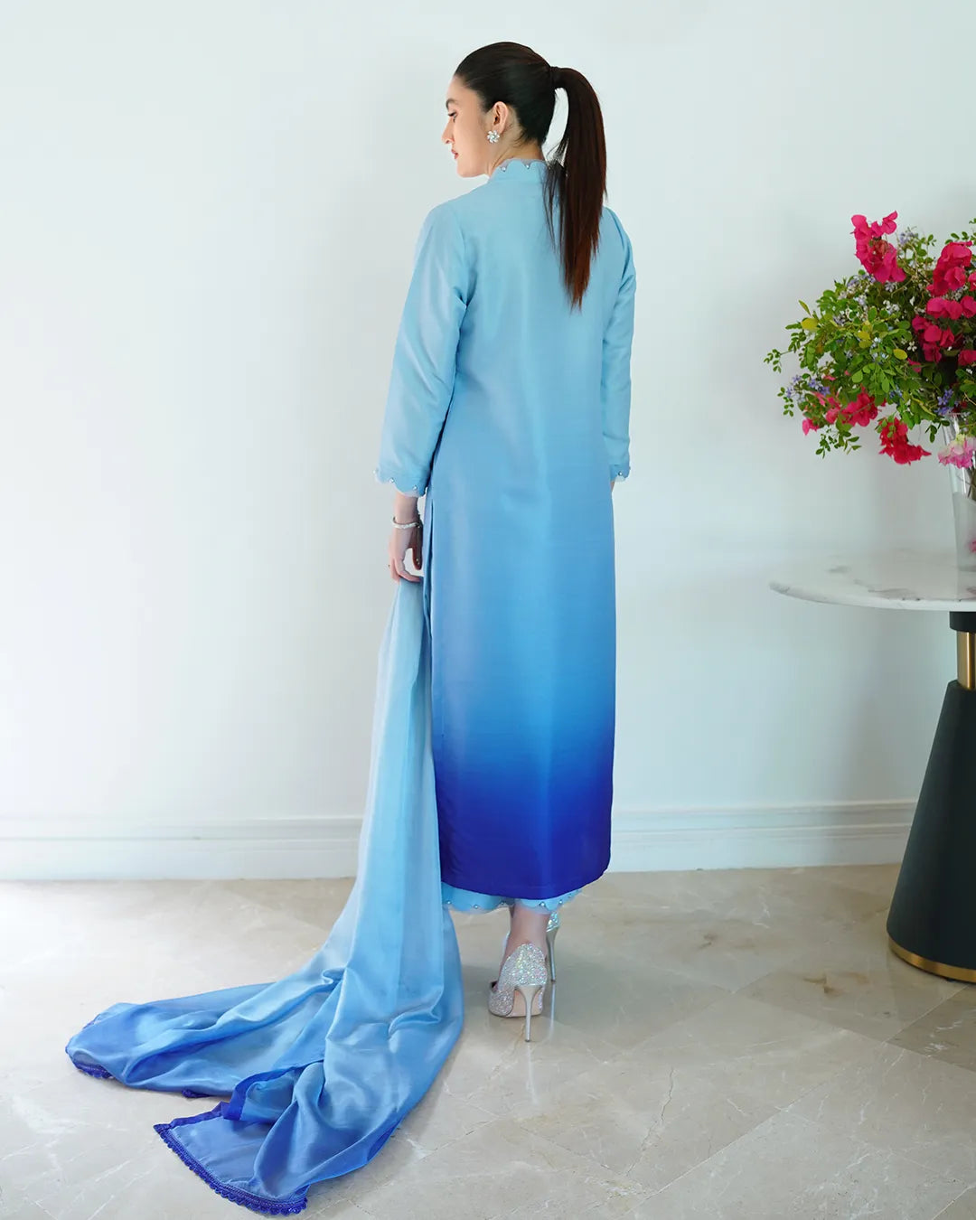 Woman wearing a blue gradient dress standing in a room with a white wall and a table with flowers.