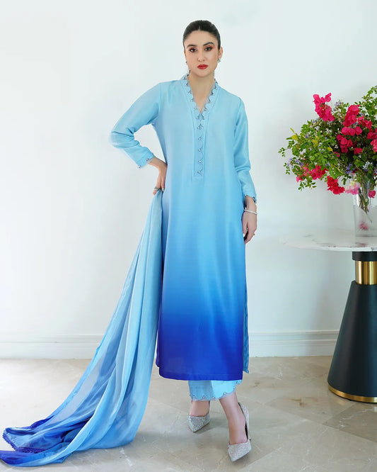 Woman in a light blue dress with a flowing fabric, standing indoors with a white wall and floral arrangement in the background.