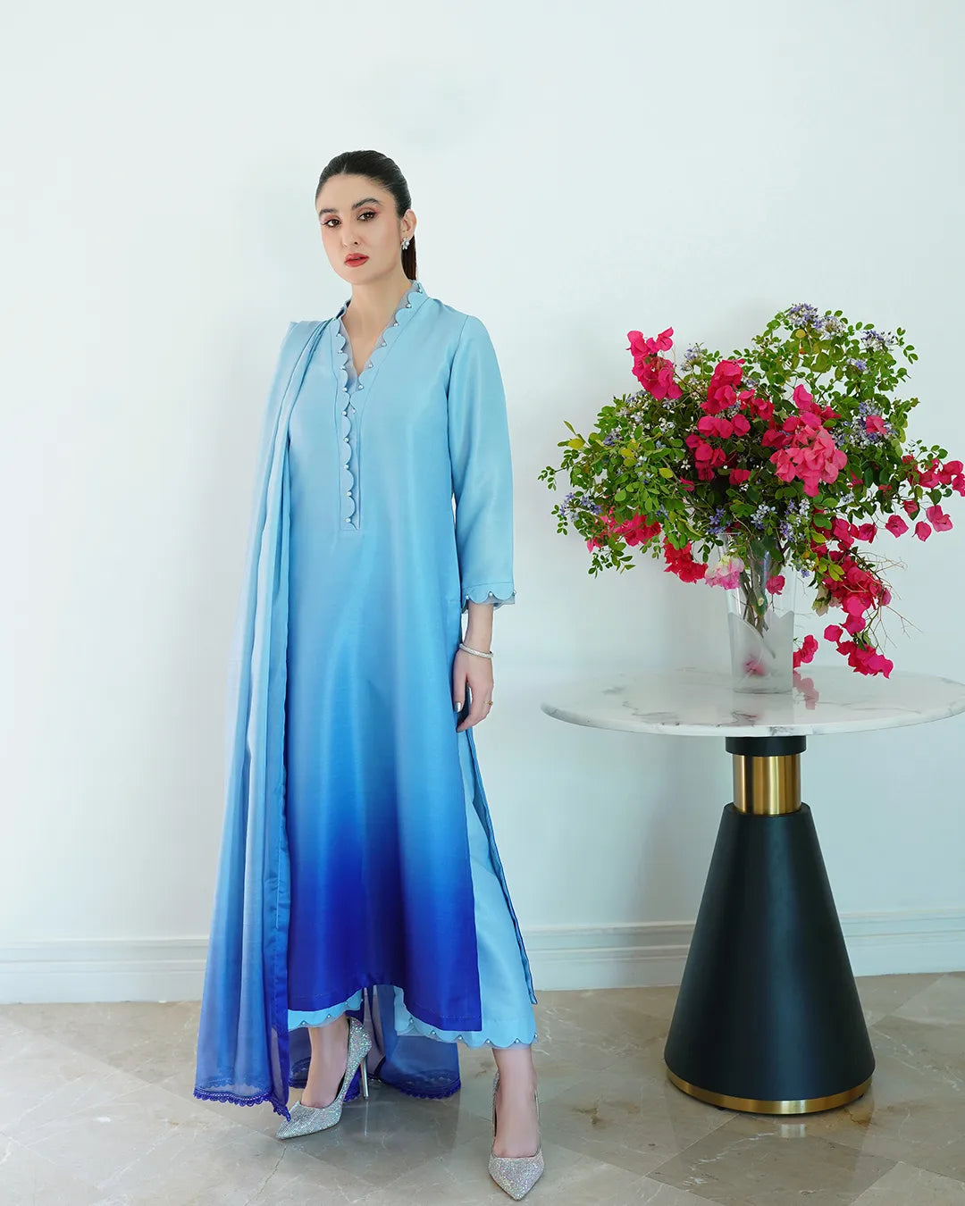 Woman in a blue gradient dress standing next to a table with flowers on a light background
