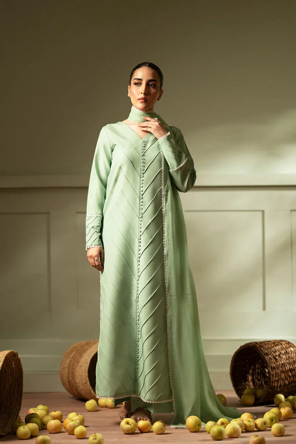 Woman wearing a light green dress standing in front of a neutral background with apples on the floor.