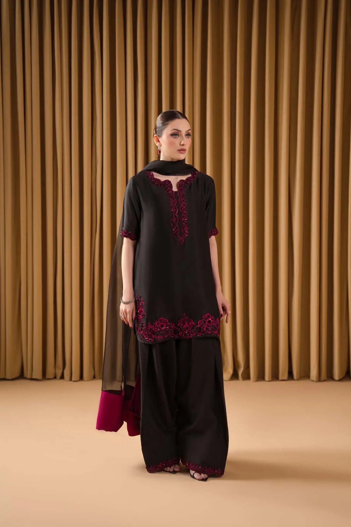Woman wearing a black outfit with red embroidery against a beige curtain background