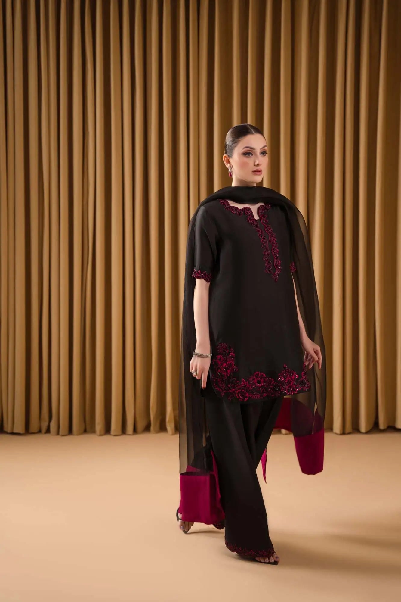 Woman wearing a black outfit with red embroidery against a beige curtain background
