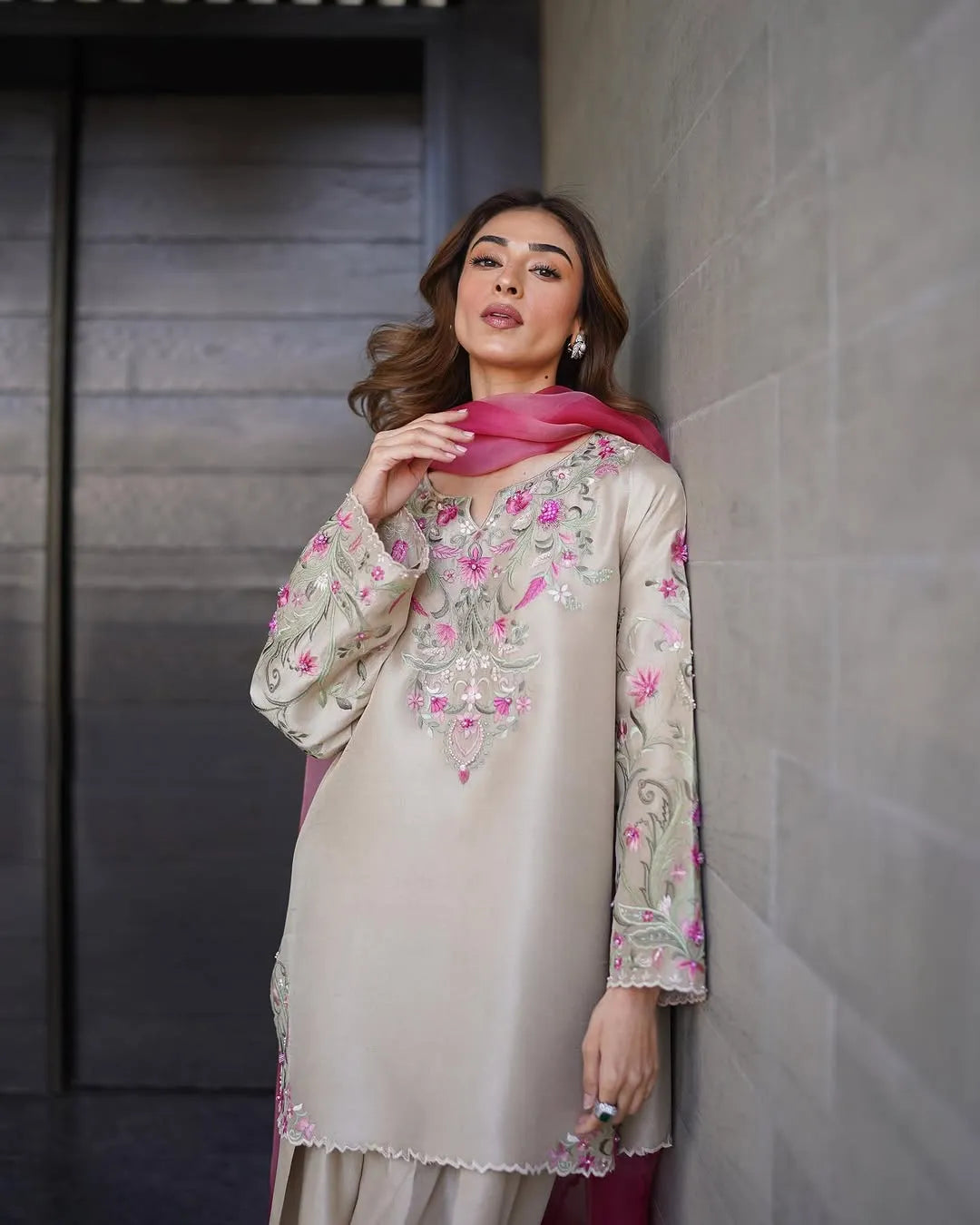 Woman wearing a beige embroidered outfit with pink accents against a gray wall.