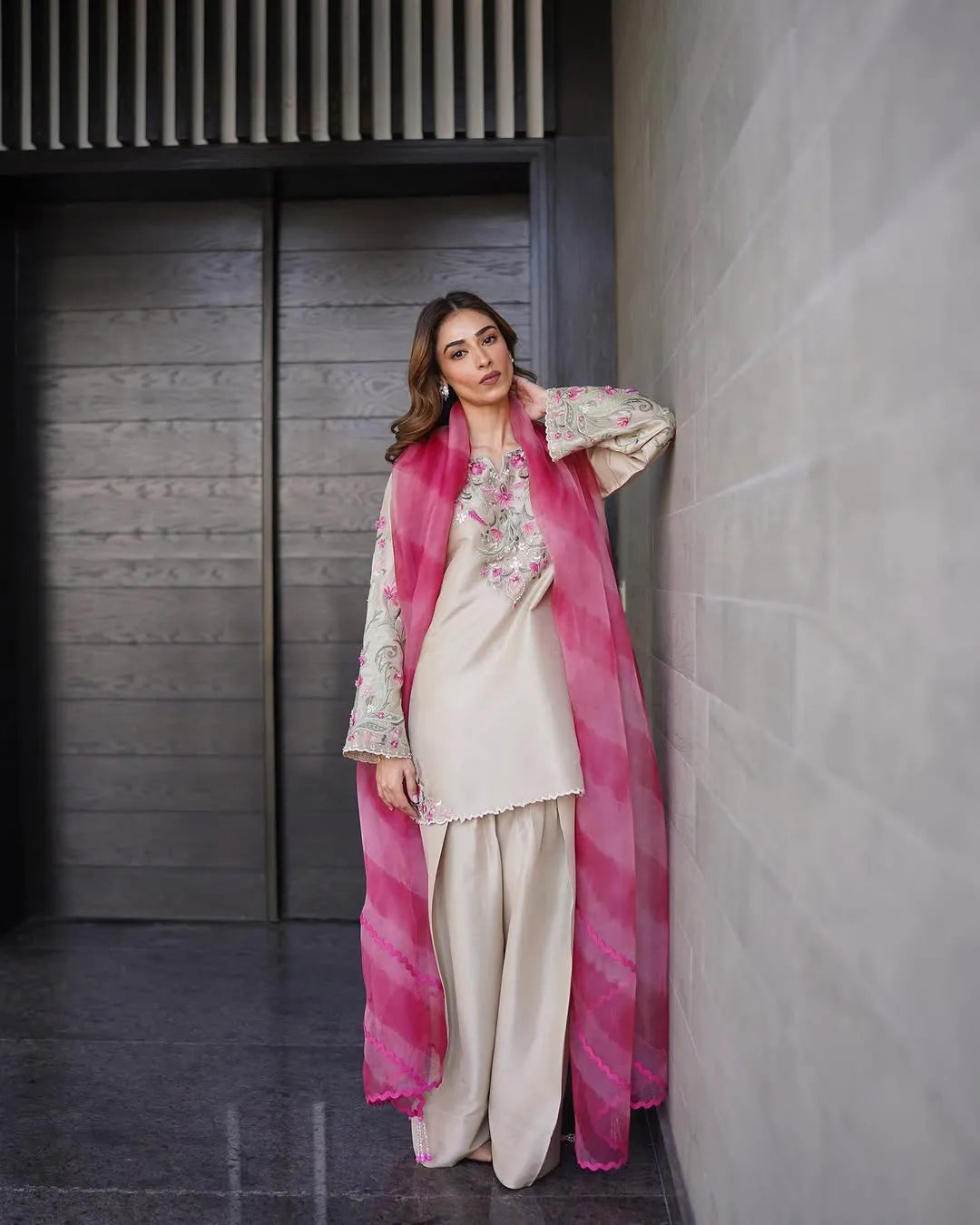 Woman in a traditional outfit with a pink dupatta leaning against a wall.