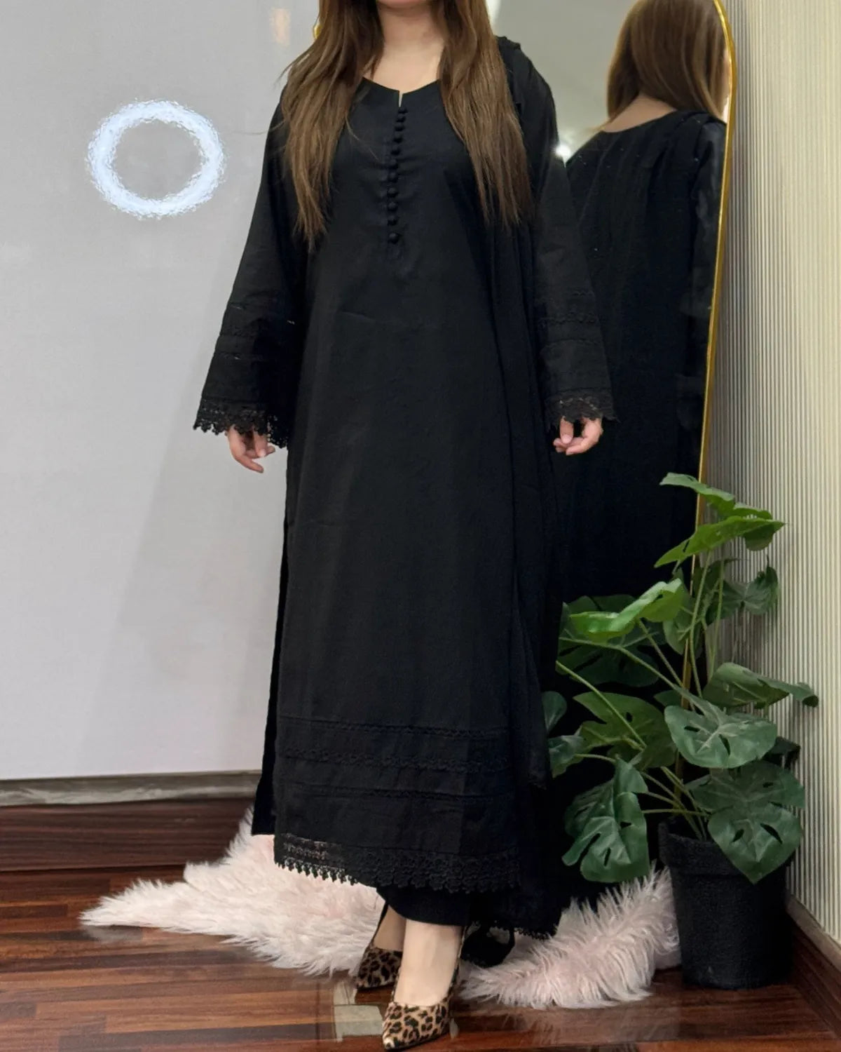 Person wearing a long black dress standing in front of a mirror with a plant in the background.