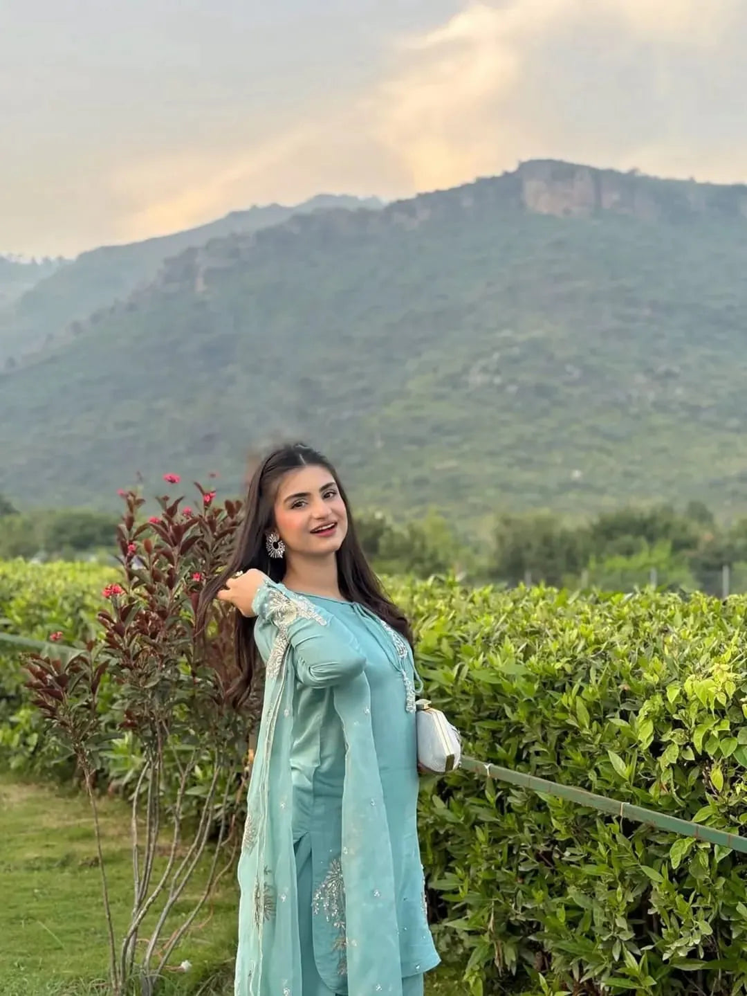Woman in a light blue outfit standing in a scenic outdoor setting with mountains and greenery.