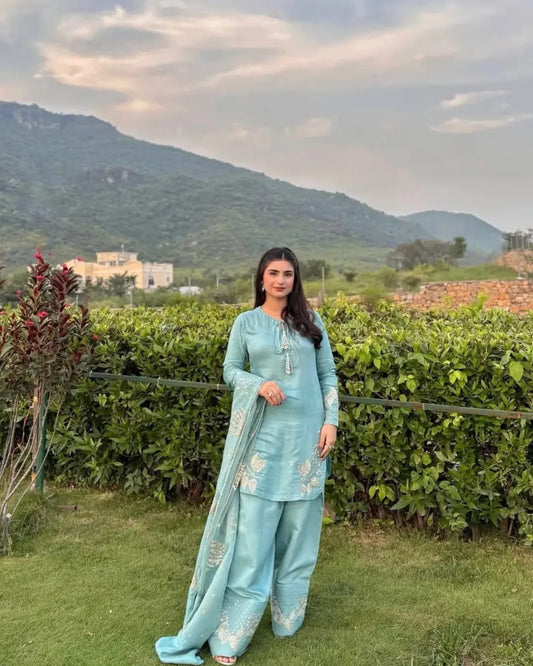 Woman in a light blue traditional outfit standing outdoors with greenery and mountains in the background