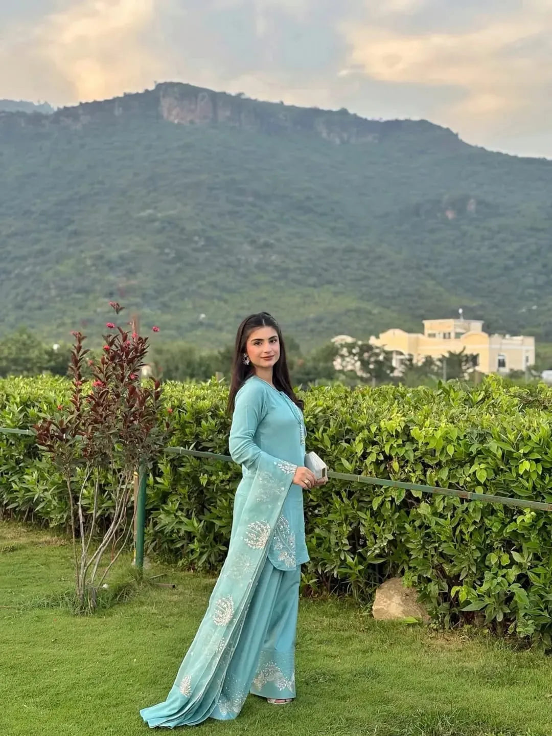 Woman in a light blue traditional outfit standing in a lush green garden with mountains in the background.