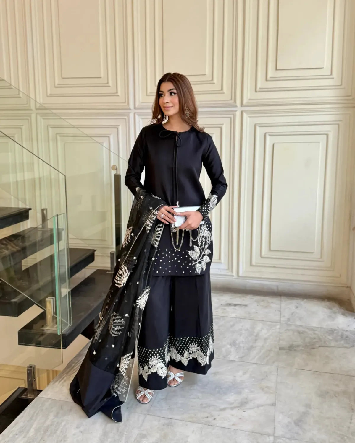 Woman in a black outfit with floral patterns standing in an elegant interior setting.