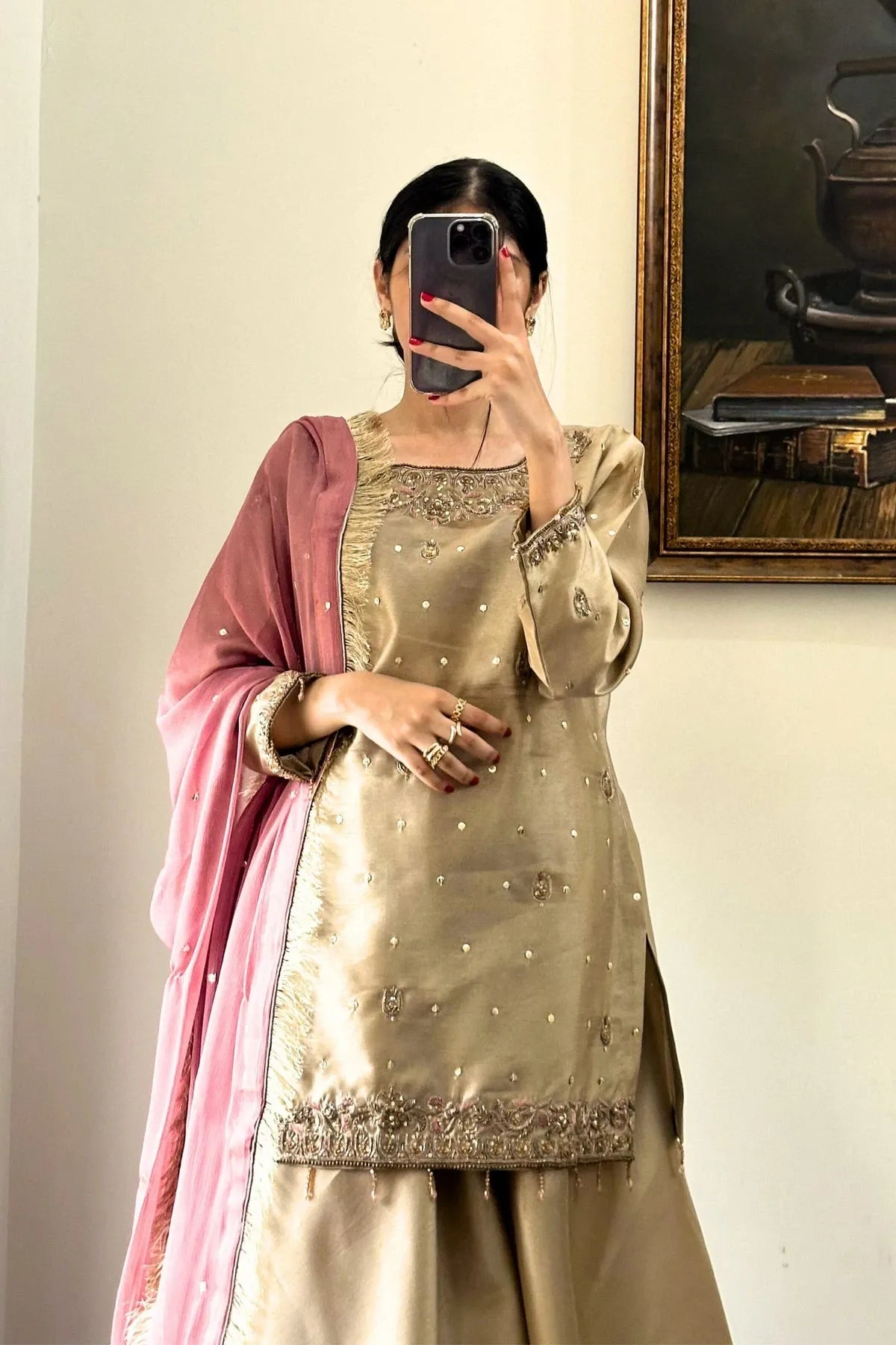 Person wearing a traditional outfit with a gold top and pink dupatta, taking a mirror selfie.