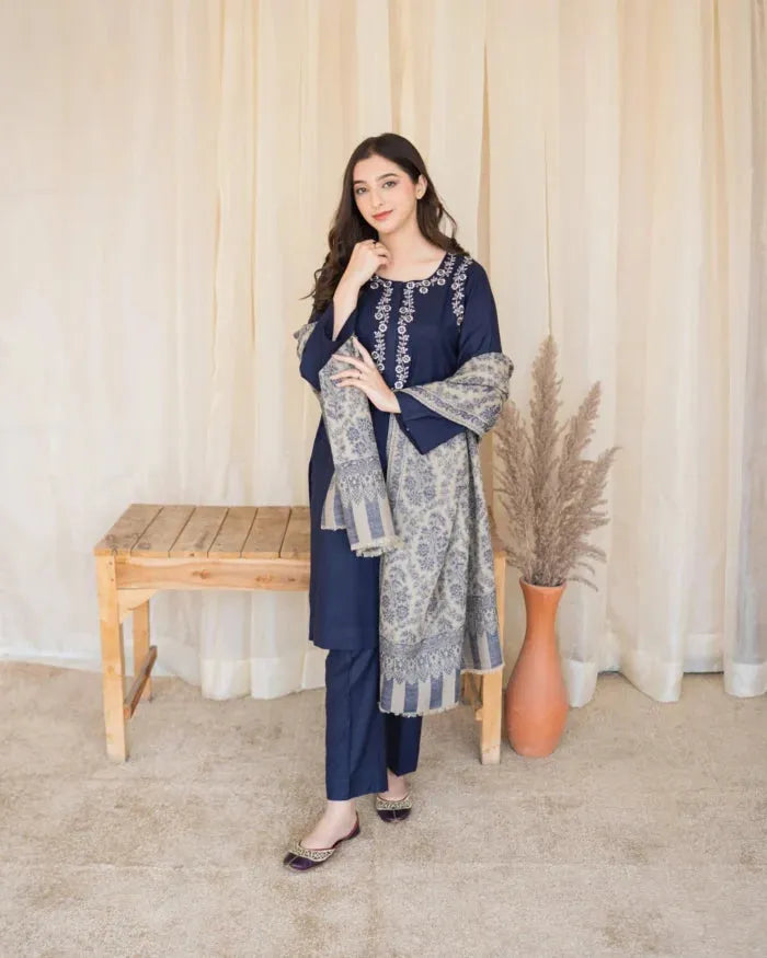 Woman in a blue and gray traditional outfit standing in a room with a wooden bench and decorative vase.