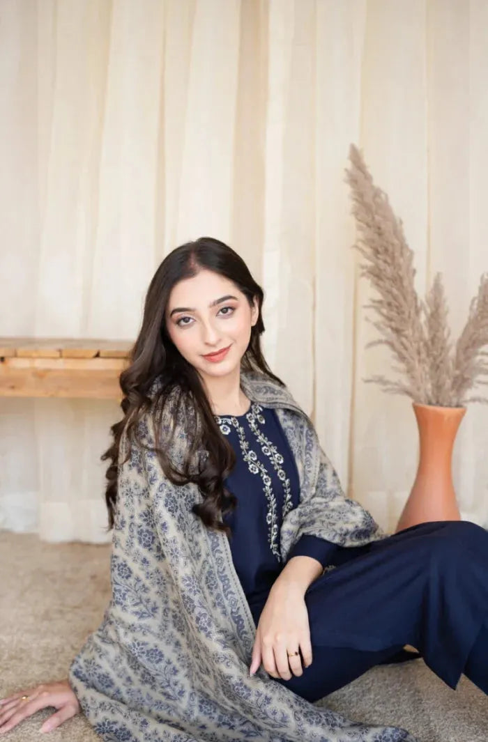Woman sitting on the floor wearing a floral shawl and dark blue outfit with a neutral background