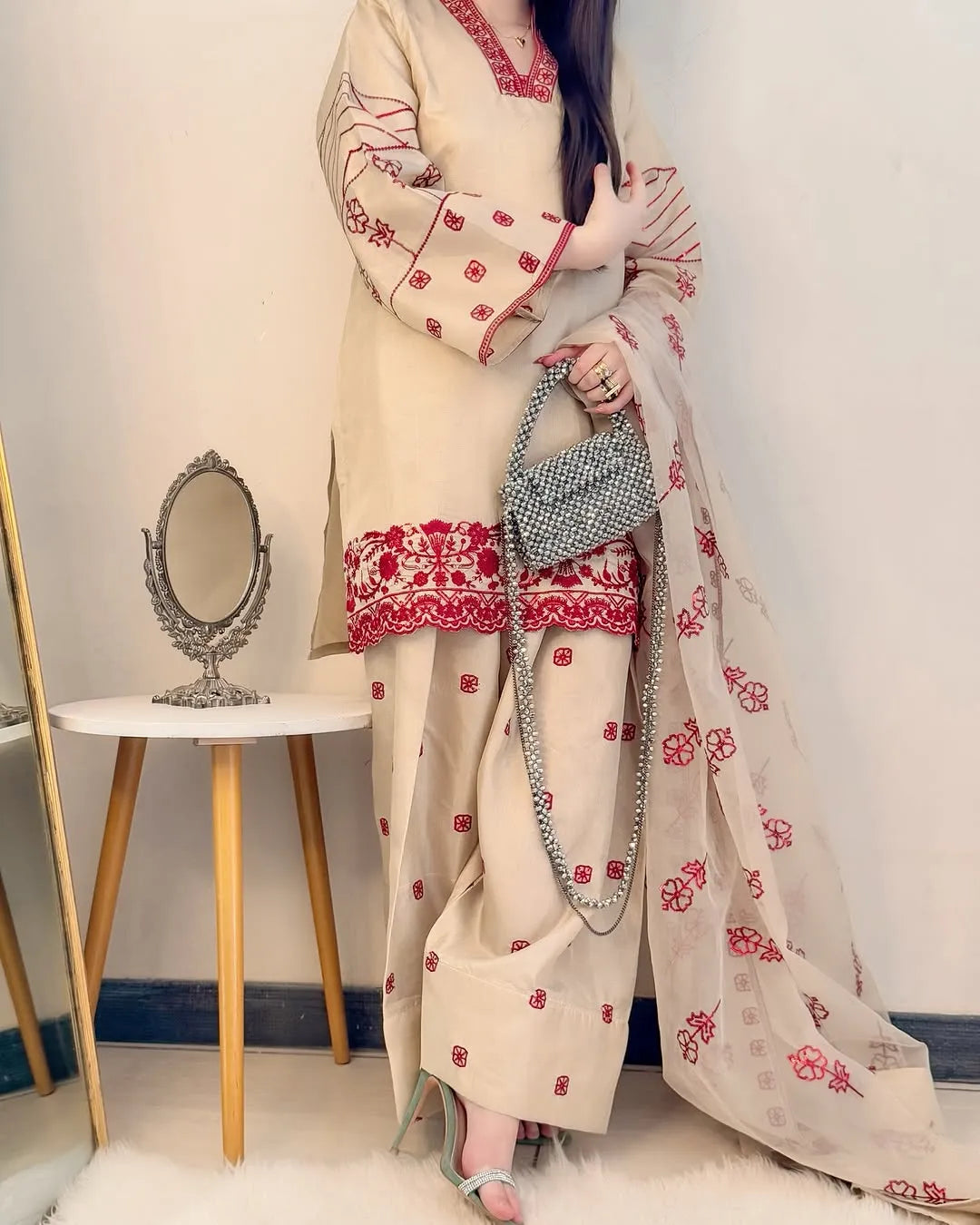 Woman in a cream and red embroidered traditional outfit holding a handbag in a minimalistic room.