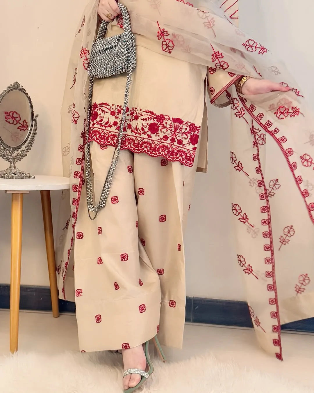 Person wearing a beige outfit with red embroidery, holding a silver handbag, in a room setting.