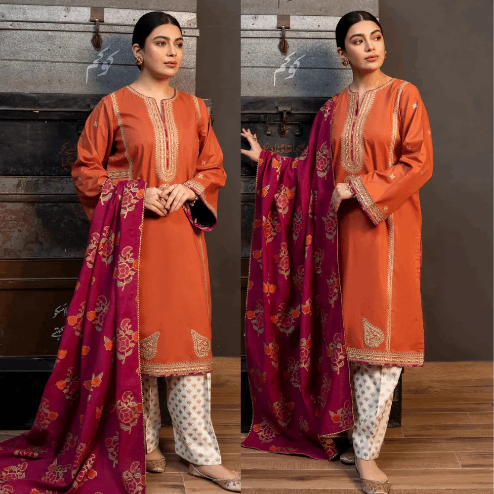 Woman wearing an orange kurta with white pants and a pink dupatta with floral patterns, standing against a wooden floor.