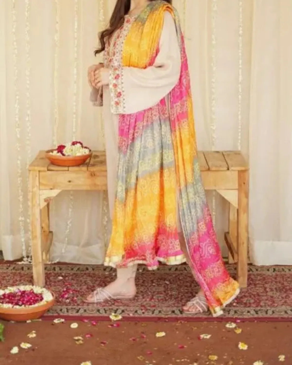 Woman in a colorful traditional outfit standing on a patterned rug with a wooden table in the background.