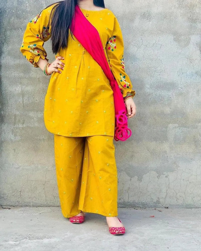Person wearing a yellow outfit with floral patterns and a pink scarf against a gray wall.
