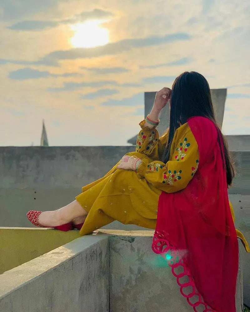 Woman in yellow and red traditional outfit sitting on a rooftop with sunset in the background