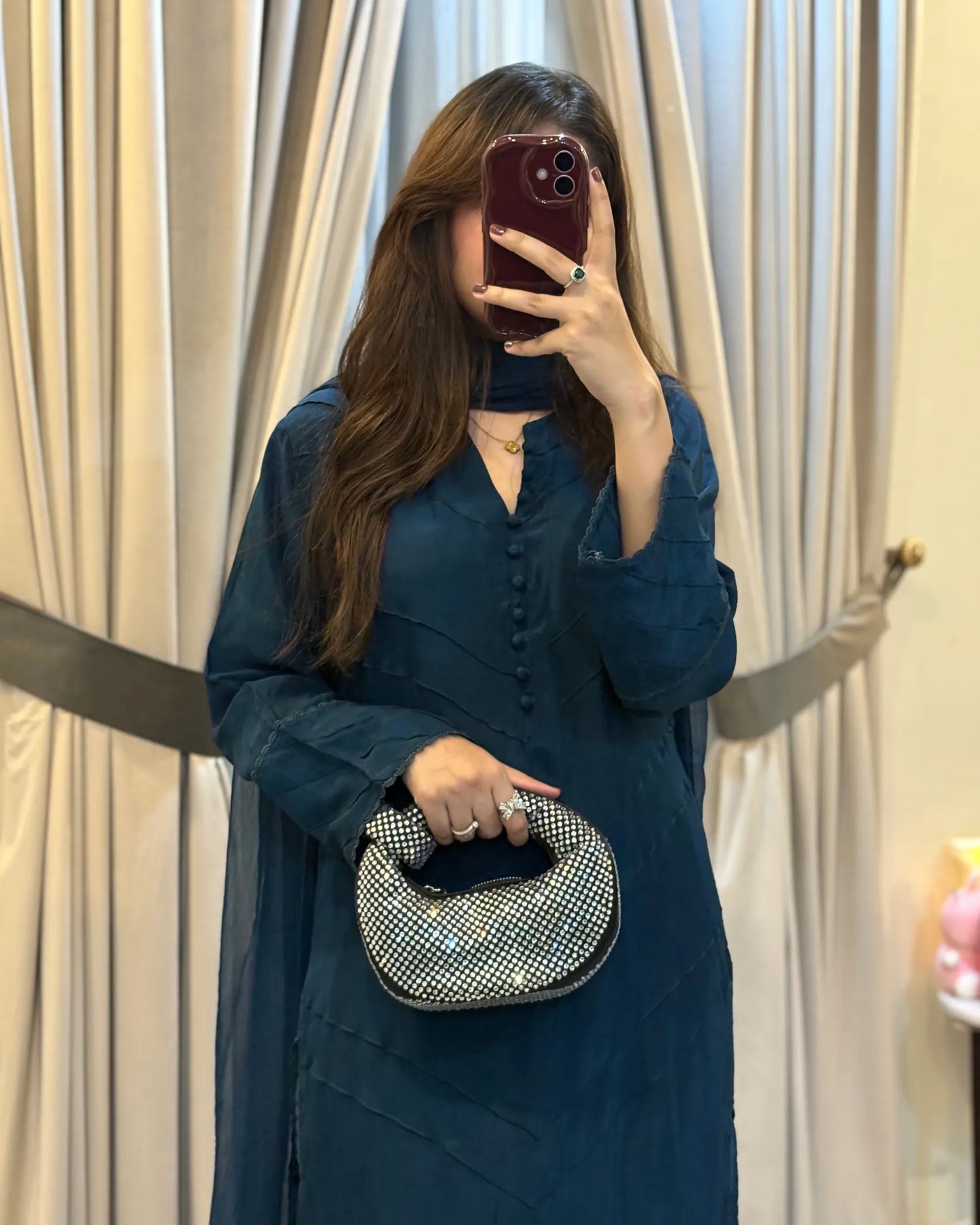 Woman in a teal dress holding a silver clutch in front of beige curtains.