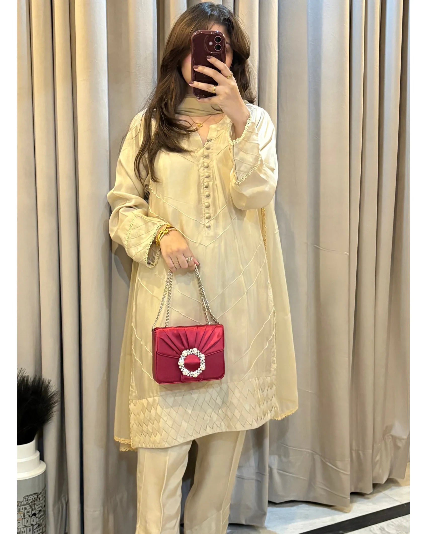 Person wearing a light-colored outfit with a pink handbag in front of beige curtains.