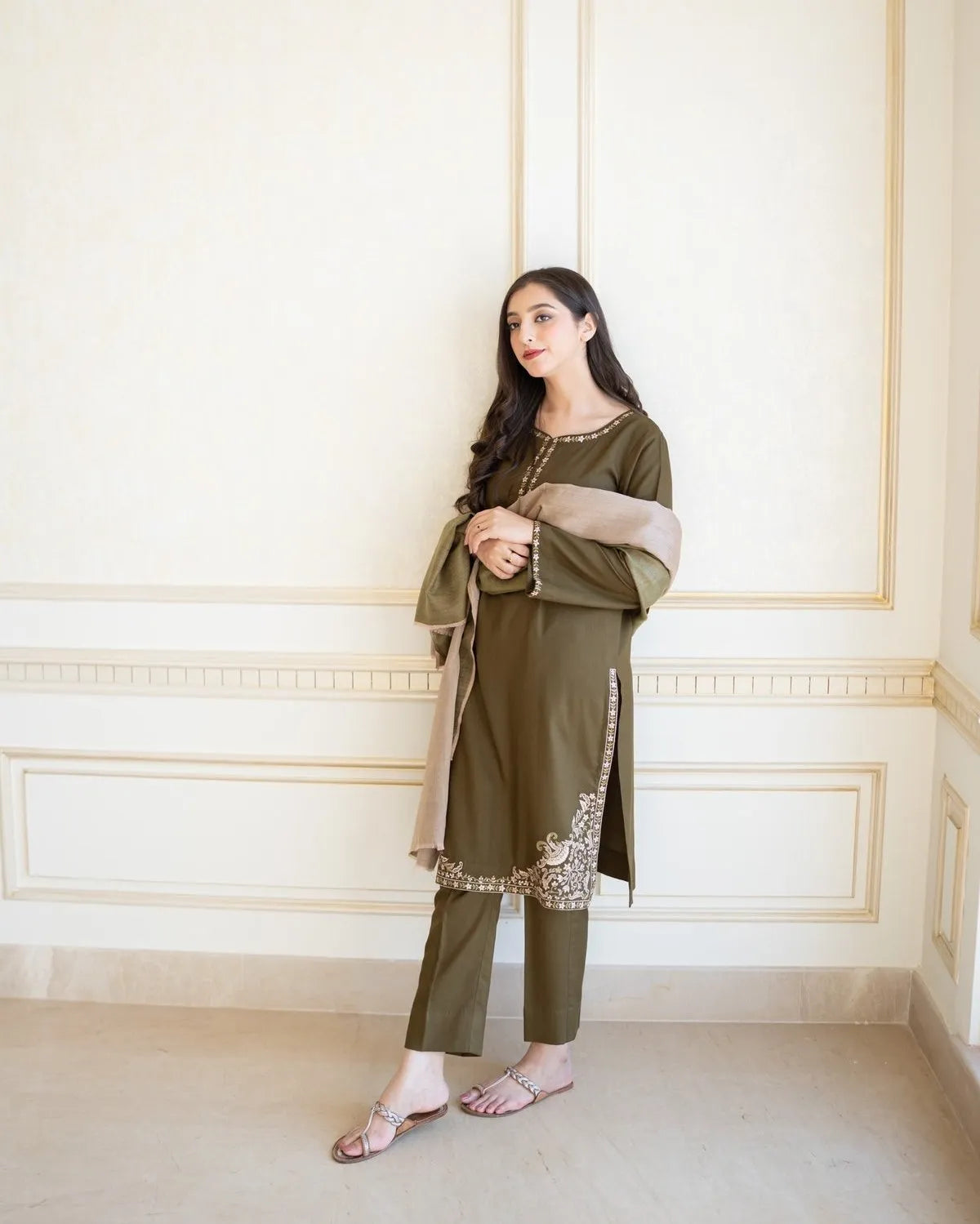 Woman in an olive green traditional outfit with a beige scarf against a white wall.