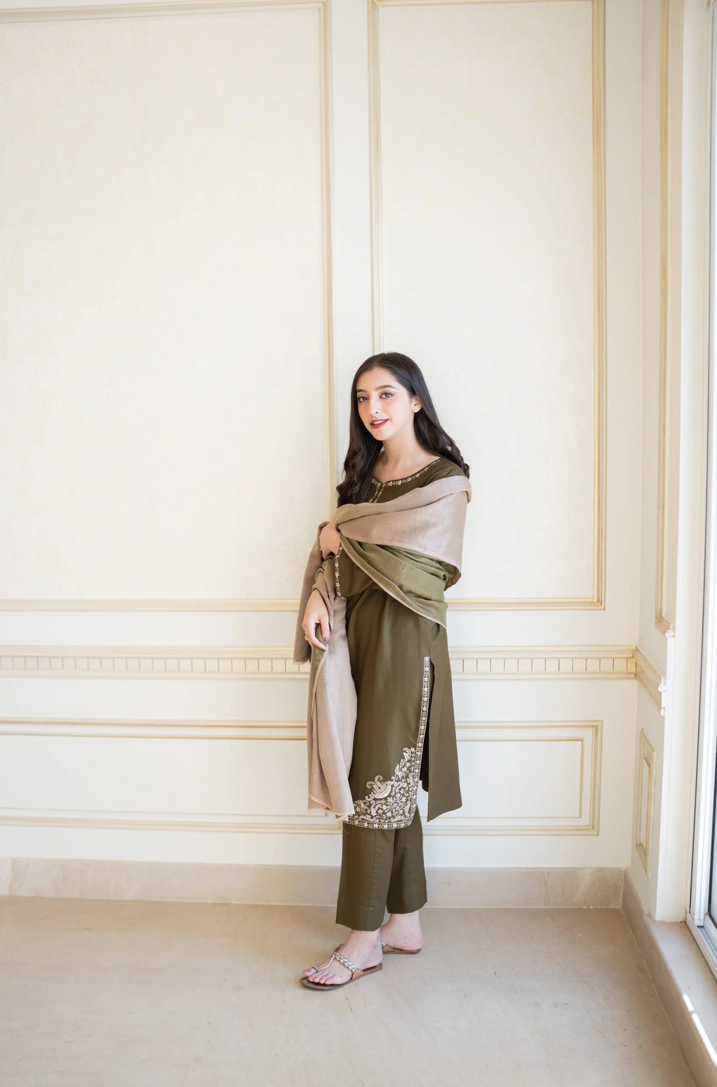 Woman in a green traditional outfit with a beige shawl against a white paneled wall.