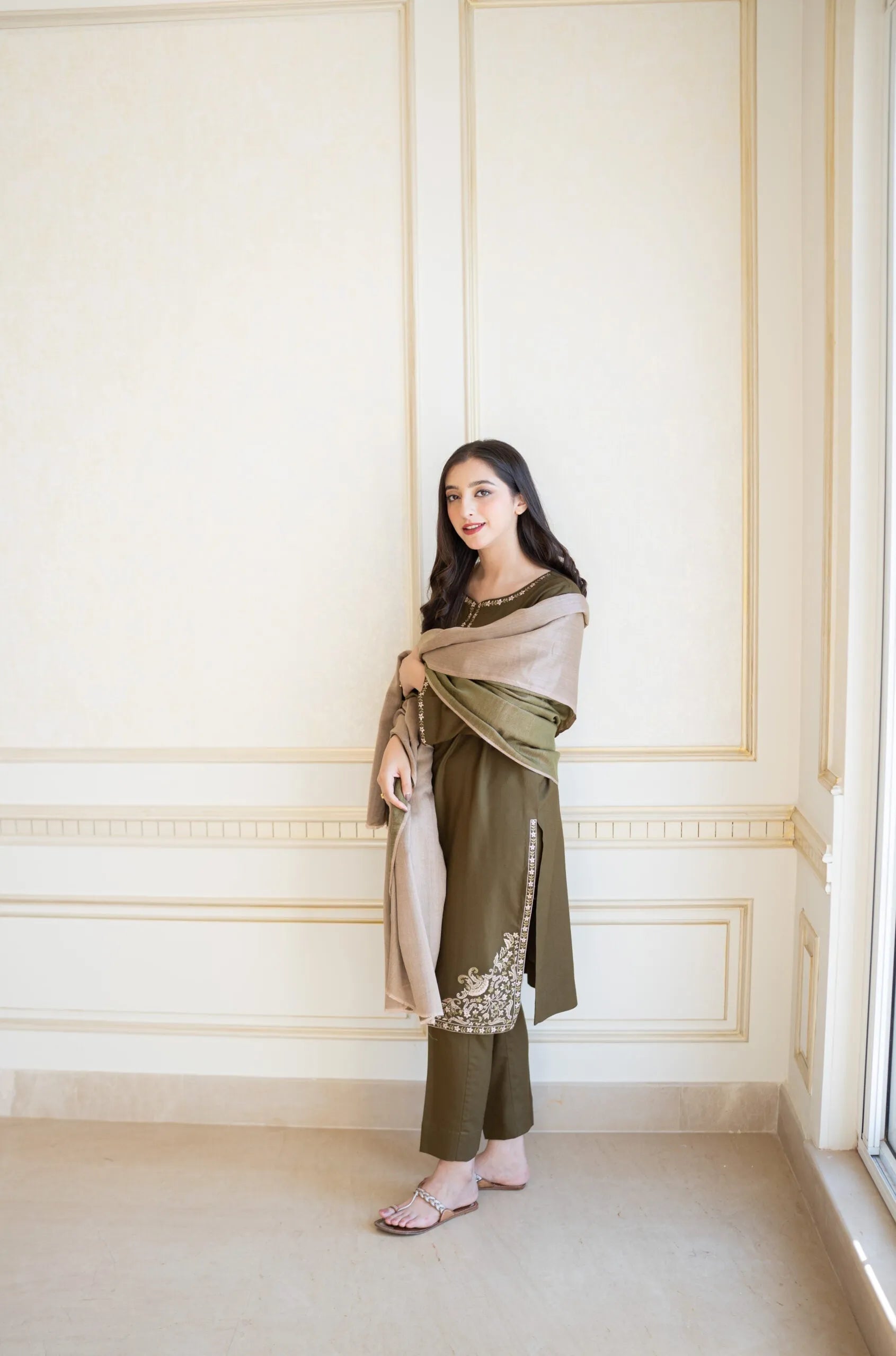 Woman in a green traditional outfit with a beige shawl against a white paneled wall.