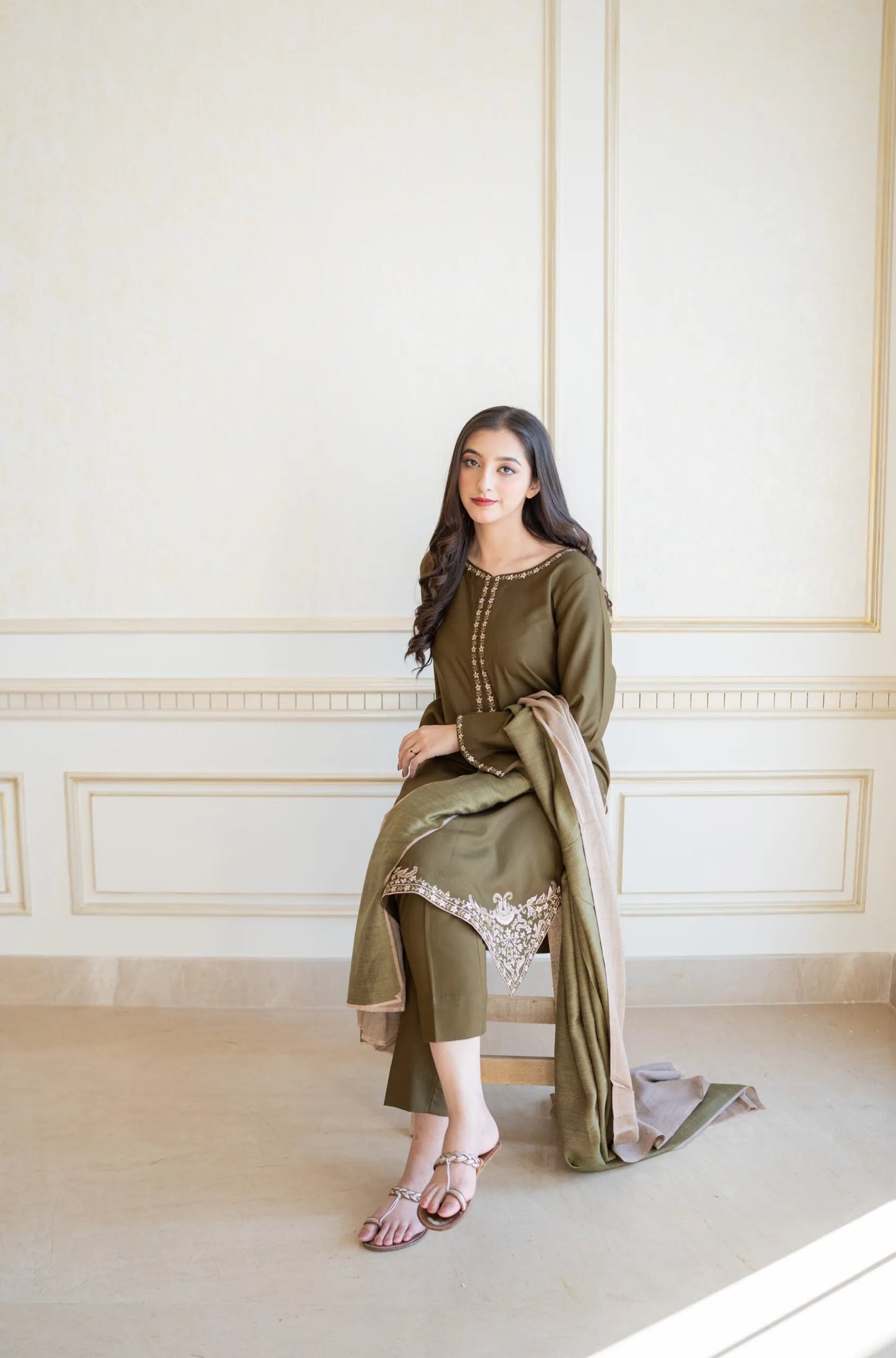 Woman in a green traditional outfit sitting on a white bench in a room with white walls.